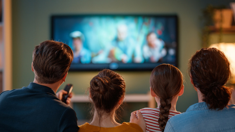 A family of four are seen sitting together on a couch and watching TV.