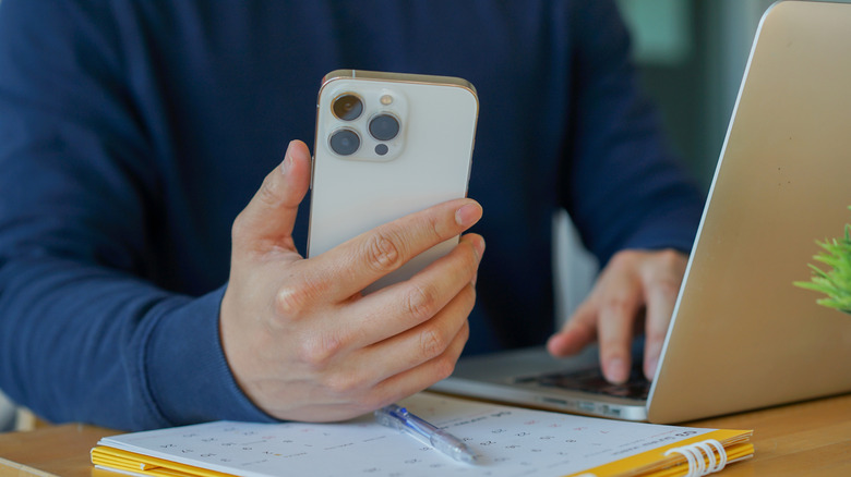 A hand holding an iPhone as it rests on a calendar on a desk, the person's other hand is on a laptop keyboard