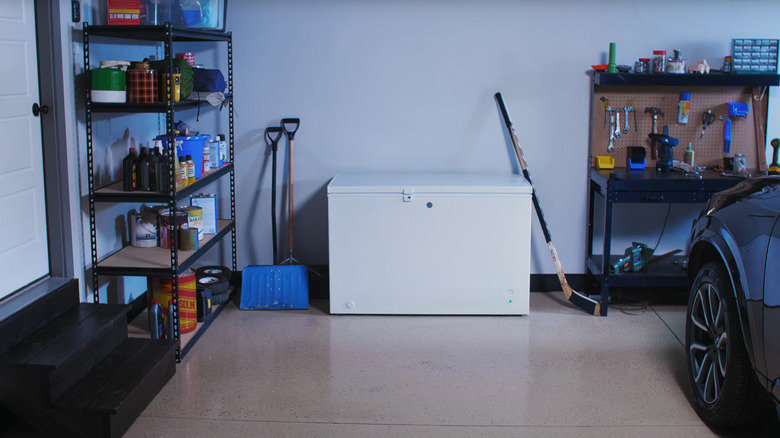 A GE chest cooler sits besides a black car, between two work shelves and next to a hockey stick and snow shovels, denoting winter time.