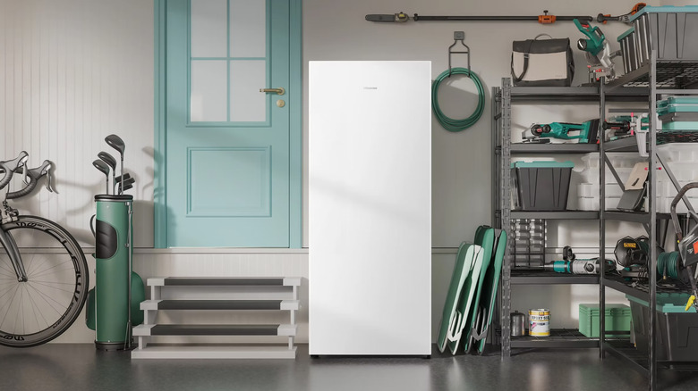A white, upright Hisense freezer sits next to a turquoise garage door, surrounded by green colored tools and golf clubs.