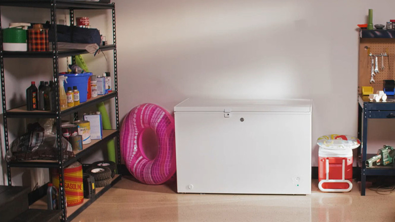 A white GE chest freezer sits between a pink donut-shaped floatie and a red cooler in a crowded garage, presumably during summer.