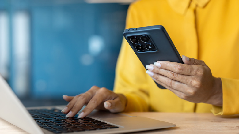 A person typing on a laptop and holding a smartphone