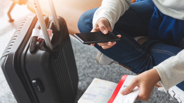 A person seated while holding a cellphone in one hand and a passport and plane ticket in the other, with a black luggage bag next to them