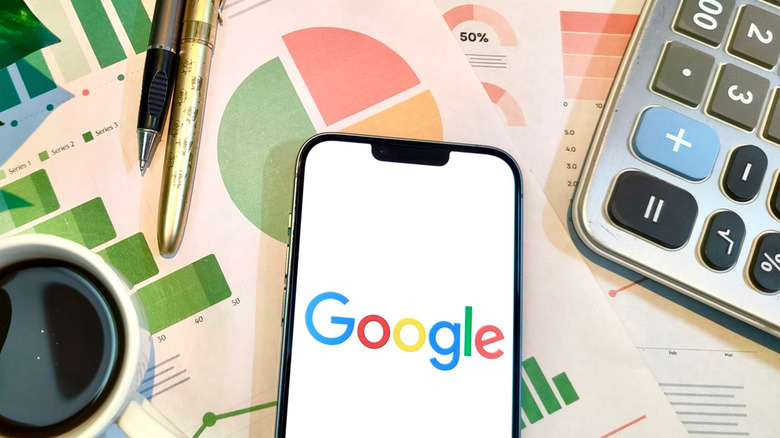 A cellphone displaying the Google logo sits atop printed graphs, while a calculator, two pens, and a coffee cup are next to the phone