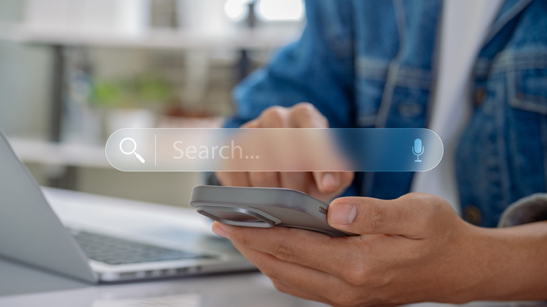 A person holding a cell phone in their left and hand and pointing at it with their right, a search bar resembling Google's is superimposed overtop of the cellphone