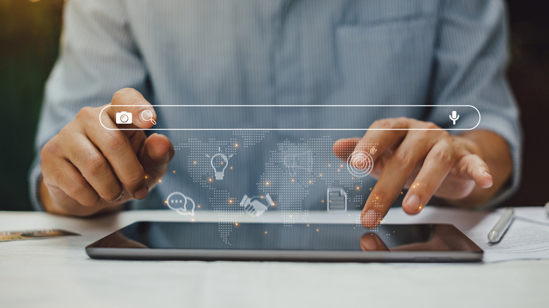 Two hands interacting with a smart tablet that is laying on a table, above the tablet is a rendered image of a search bar and several different icons