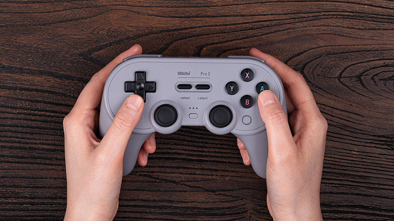 Hands holding a gray 8BitDo Pro 2 gaming controller against a dark wood background