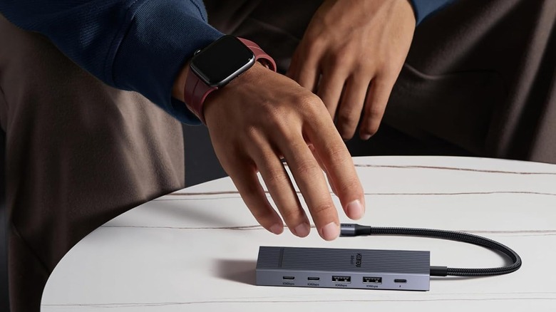 A USB hub sitting on a white table with the hand of someone wearing an Apple Watch reaching for it