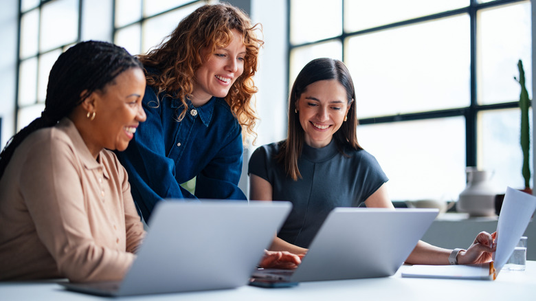 Three women in an office smiling in front of laptops.