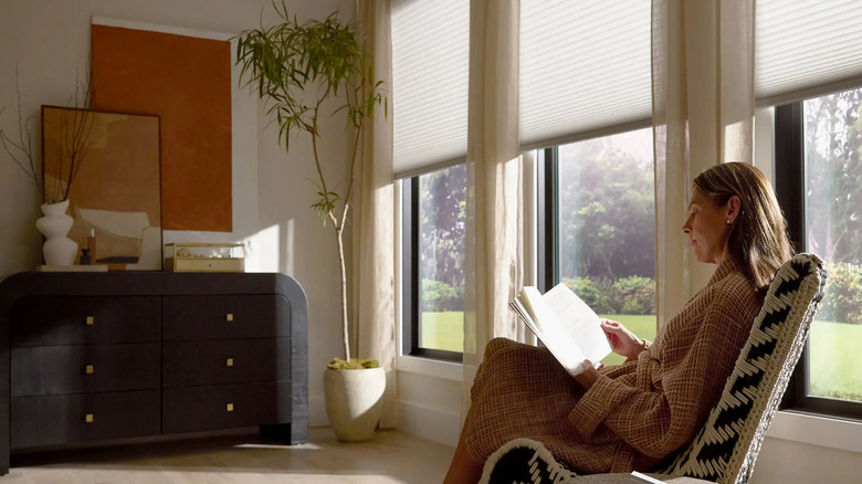 A woman reads a book in a chair, her Serena smart shades cover half of her windows