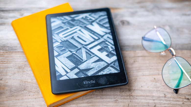 Kindle e-reader on a yellow book beside eyeglasses on a wooden table.