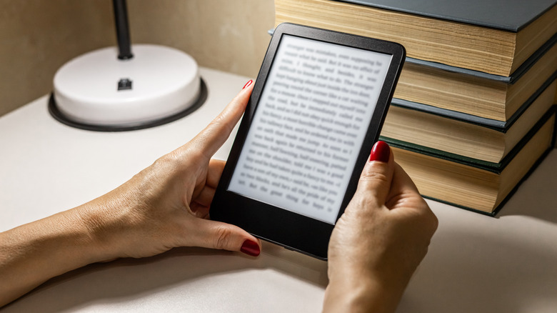 Person holding an e-reader beside a stack of books.