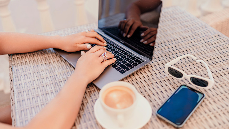A person using a MacBook with a cup of coffee, glasses, and iPhone placed next