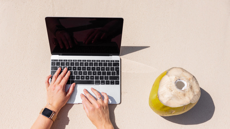 Woman using a laptop on a beach next to a coconut