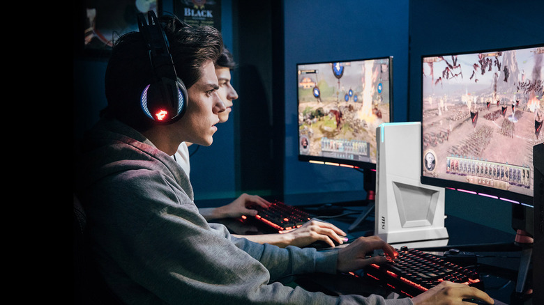 Two men play a PC game side-by-side in a darkly lit room