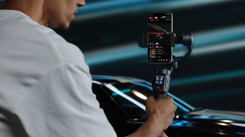 A person in a white shirt holds the Hohem iSteady M7 AI Tracking Smartphone Gimbal in their right hand as they take photographs of a fancy black car