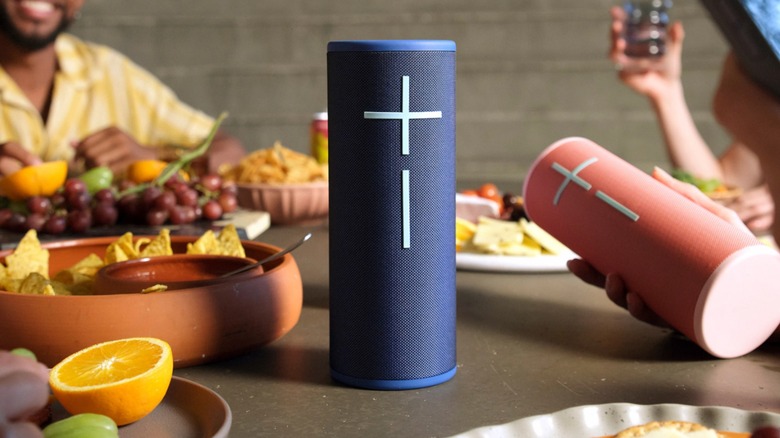 A blue Ultimate Ears Megaboom 4 Bluetooth speaker sits on a table as a cropped hand holds a red version of the speaker