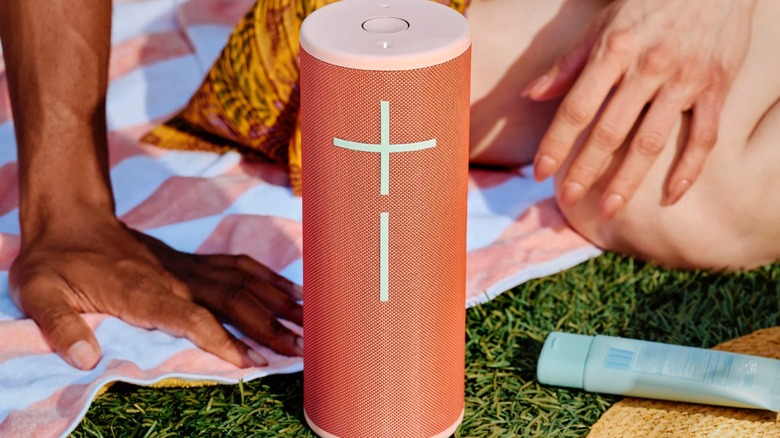 A closeup of a red Ultimate Ears Megaboom 4 Portable Bluetooth Speaker, the speaker itself is laying on some grass at what appears to be a picnic