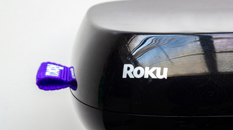 The tail-end of a black Roku Remote, the plastic case displays the logo, as does the purple material that hangs from the end of the remote