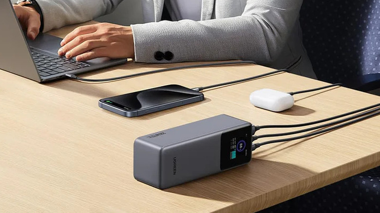 A UGREEN Nexode power bank with a digital display on a table with cords attached to a computer, a smartphone, and an AirPods case