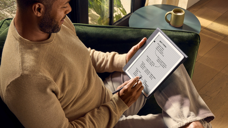 Man writing notes on a Kindle Scribe with a stylus while sitting on a couch.
