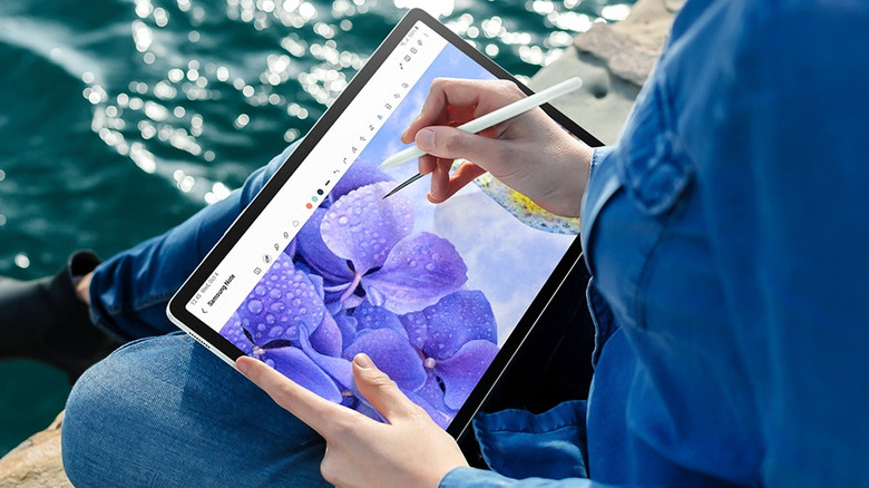 Person drawing a purple flower on a Samsung Galaxy Tab S9 FE with an S Pen outdoors by the water.