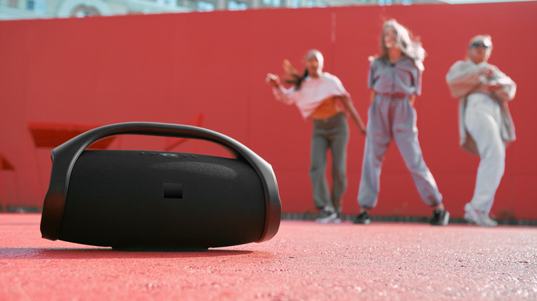 A wireless speaker placed on the ground with a group of people dancing in the background