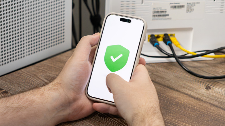 Person's hand holding a smartphone with a checkmark and shield onscreen, in front of a router
