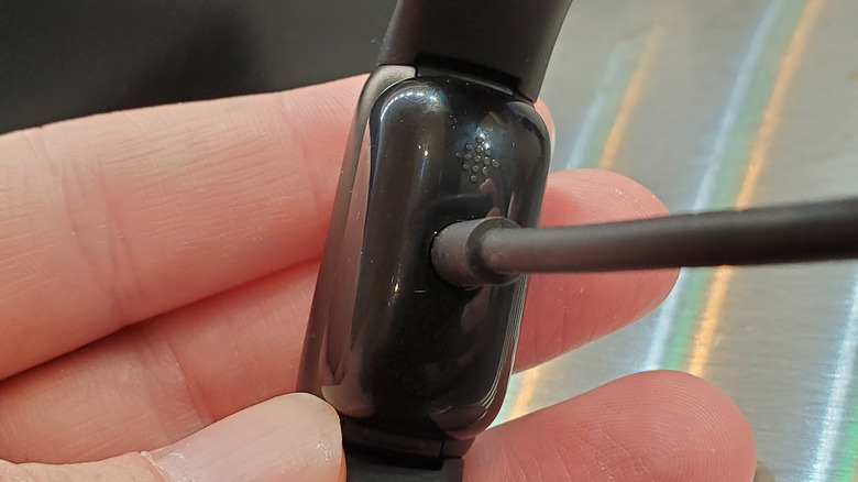 A hand holding a Fitbit Luxe, showing the back attached to its charging cable.
