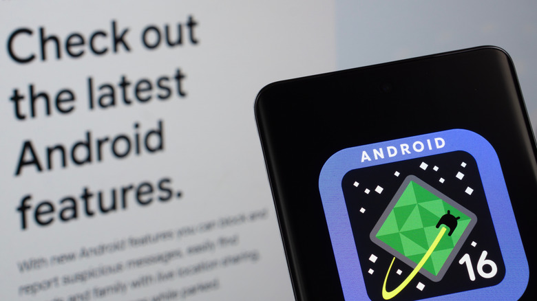 smartphone showing android 16 logo with "check out the latest android features" on a monitor in the background