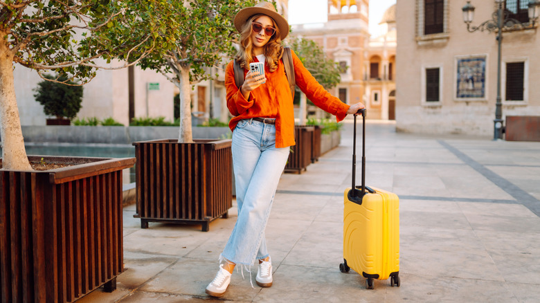 A woman standing on the streets while holding a suitcase and looking at her phone