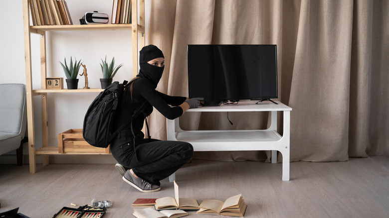 A masked burglar crouching in a living room while unplugging or stealing a flat-screen TV.