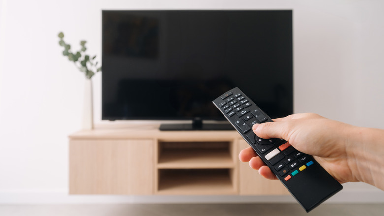 A hand holding a TV remote control pointed at a flat-screen television in a minimalist living room, with the TV and media console blurred in the background.