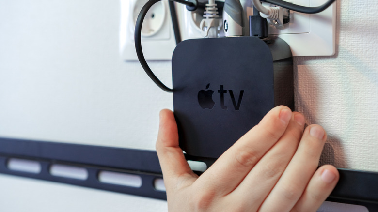 A close-up of an Apple TV streaming box being mounted or positioned behind a wall-mounted TV.