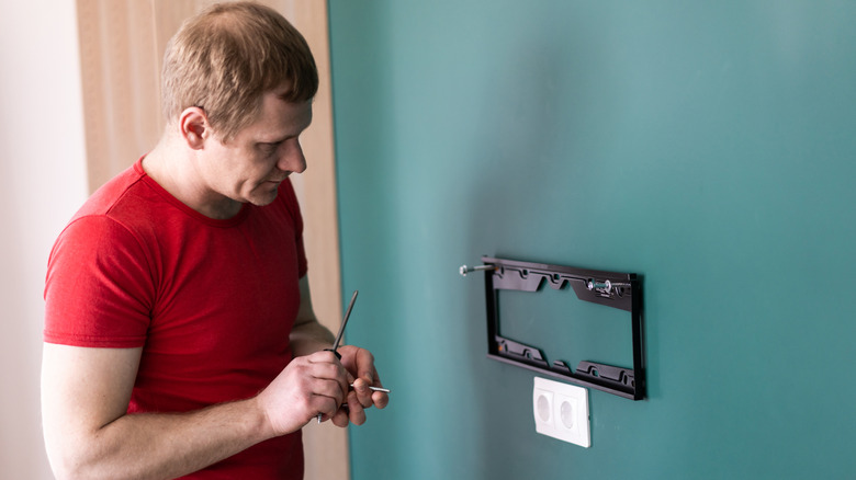 A person installing a wall-mount bracket for a flat-screen TV on a teal-colored wall.