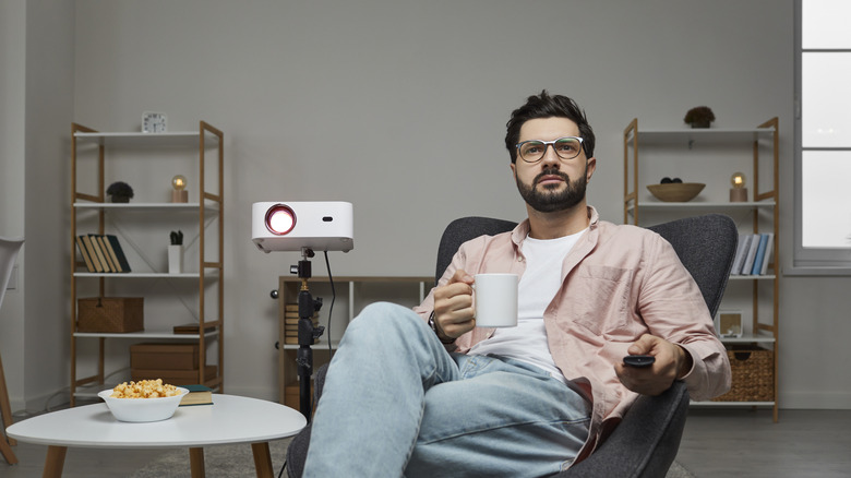 A person watching a projector in a living room while holding a remote and coffee mug.