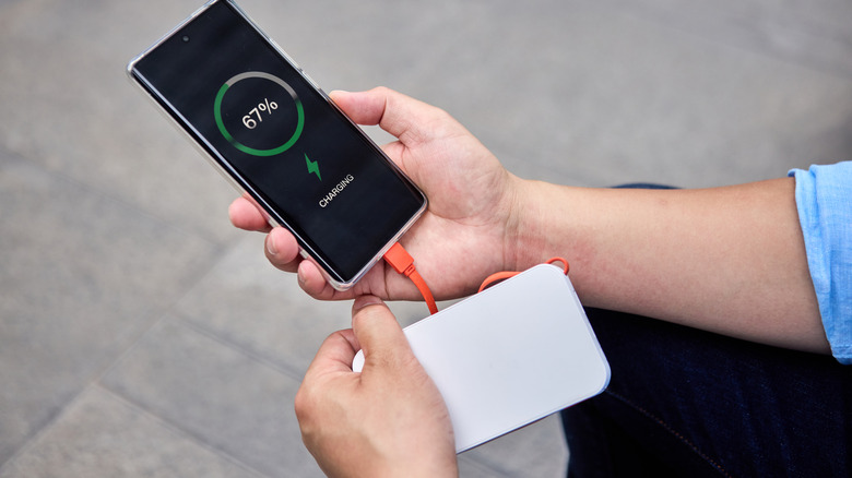 A person holding a phone and a power bank, the charging symbol on the phone screen.