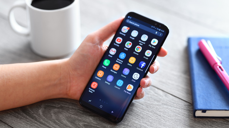 a person holding a Samsung Galaxy S9 Plus next to a mug and a notebook with a pen