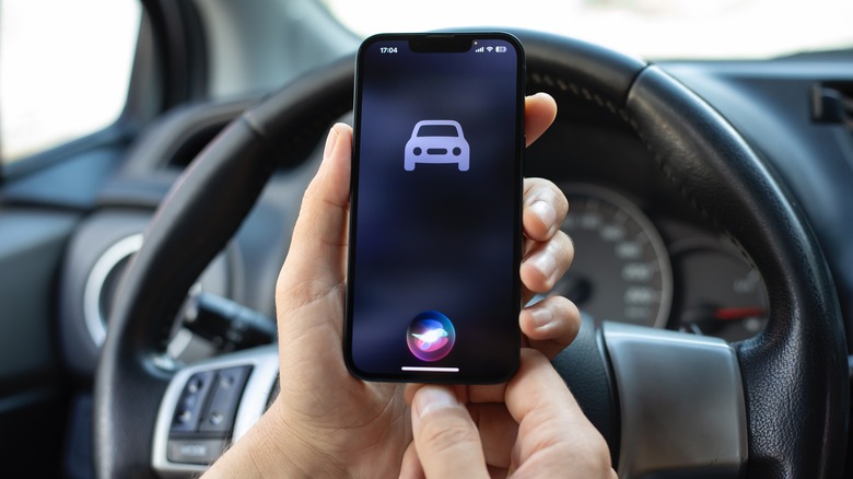 Both an individuals' hands hold an iPhone that displays a 2D rendering of a vehicle as well as the classic Siri shape, the photo is taken from the perspective of the individual as they sit behind the wheel of a vehicle