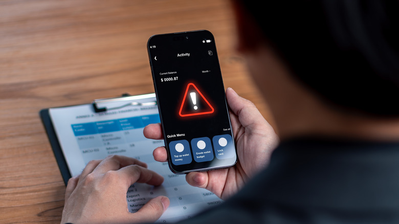 An individual looking at a smartphone, the display of the phone shows a bank account that has been maliciously attacked