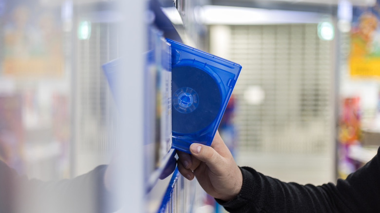 An individual's right hand is grabbing a translucent-blue disc case from a shelf, the packaging allows the disc to still be viewable inside the container