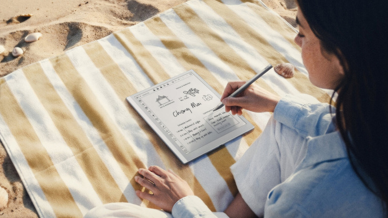 A woman on the beach writing on the Boox Go 1.03 Gen II tablet that's on a striped blanket.