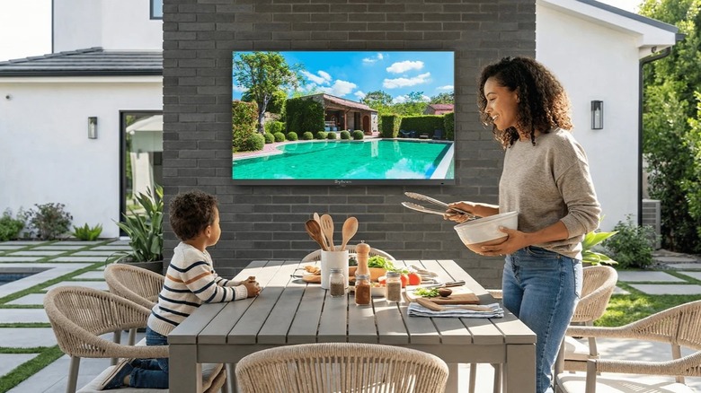 A Sylvox frameless outdoor TV mounted to a wall with a family preparing to eat in the foreground.