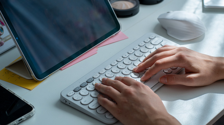 Hands typing on a white wireless keyboard beside a tablet and a wireless mouse on a desk.
