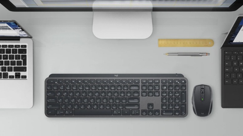 The keyboard and mouse of the Logitech MX Keys Combo on a desk, beside laptops and a monitor.