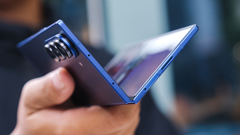 Hand holding a partially unfolded blue foldable phone with the hinge and rear cameras visible.