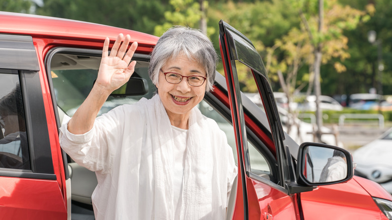 A woman waves to someone offscreen from the front passenger side of a car.