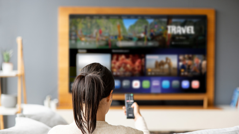 woman watching TV, blurred in background, using remote to control it