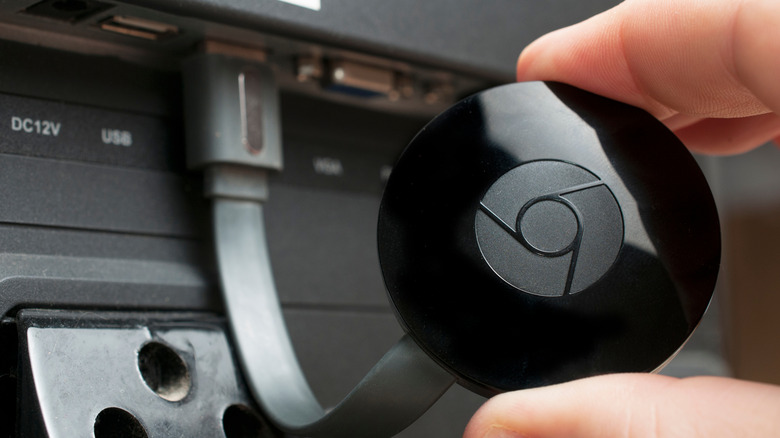 A Google Chromecast held in between the thumb and forefinger of someone's right hand, the Chromecast is connected to the HDMI port on a TV