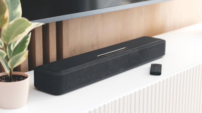 A black soundbar and remote sits atop a white TV stand, a potted plant is next to the device